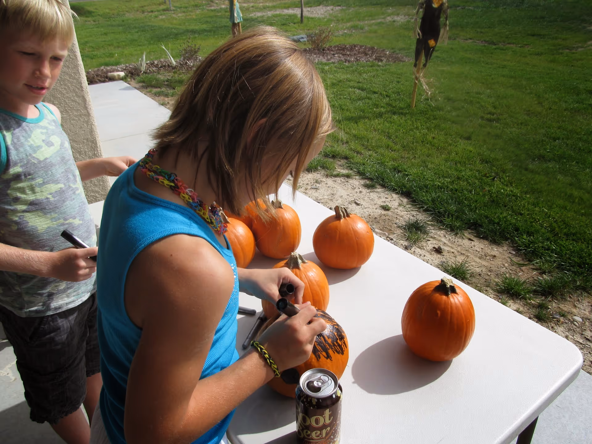 Two children outdoors at a table with small pumpkins. One child is drawing or writing on a pumpkin with a black marker, while the other child watches. There is a can of root beer on the table and green grass in the background.