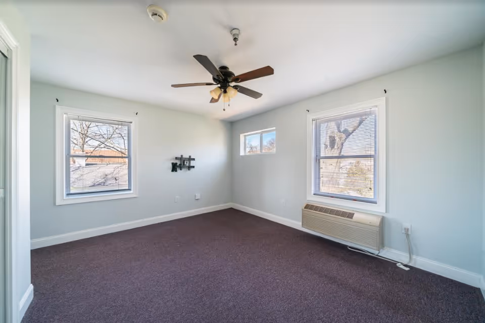 Empty carpeted bedroom with light blue walls, two windows, a ceiling fan, and a wall air-conditioning unit.