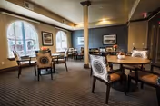A spacious dining area with several round tables and cushioned chairs featuring patterned upholstery. Large arched windows allow natural light to fill the room, and framed artwork is displayed on the walls. The room has a warm, inviting atmosphere with neutral tones and carpeted flooring.