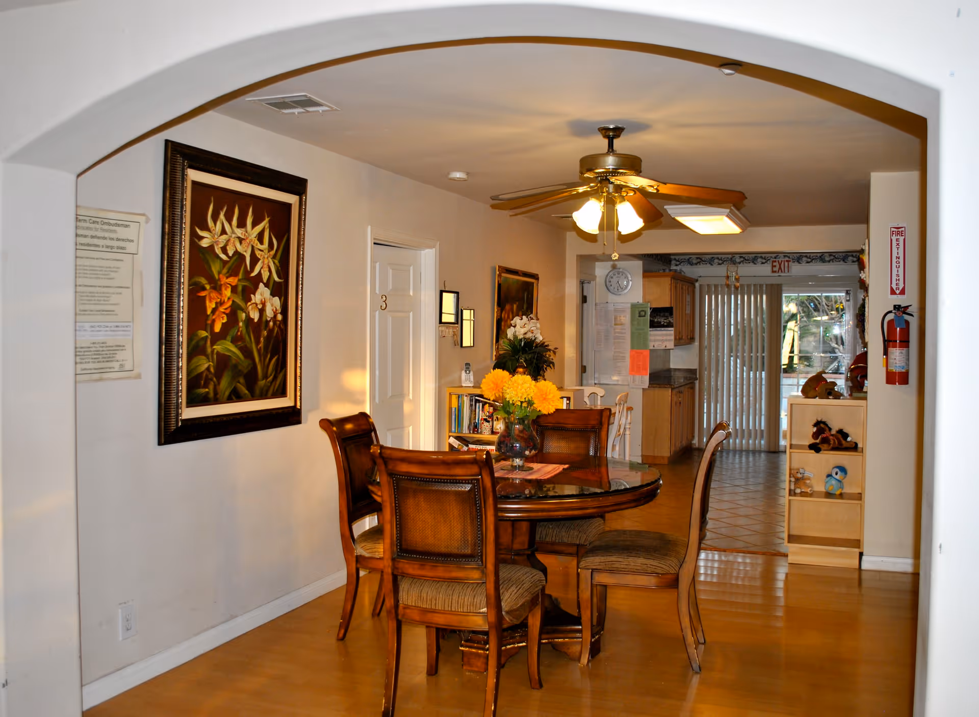 Interior view of a senior living facility dining area with a round wooden table and four chairs. A vase with yellow flowers is placed on the table. The room has light-colored walls, a ceiling fan with lights, and a framed floral painting on the wall. In the background, there is a kitchen area with wooden cabinets and a sliding glass door with vertical blinds leading outside. A fire extinguisher is mounted on the right wall near the exit sign.