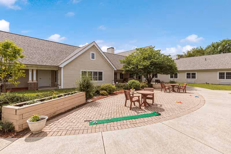 Outdoor patio area at Southpointe Healthcare Center (WI) featuring a paved brick seating area with round wooden tables and chairs, surrounded by greenery and trees, adjacent to a beige building under a partly cloudy sky.