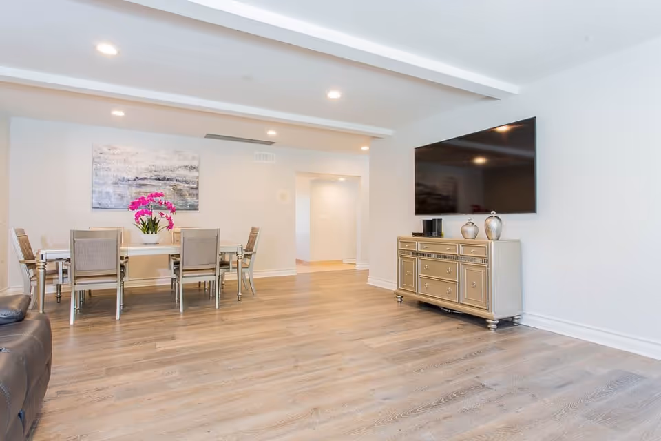 A bright and spacious interior room with light wood flooring, a dining table with six chairs and a vase of pink flowers, a large flat-screen TV mounted on the wall above a decorative cabinet, and a piece of abstract artwork on the wall.