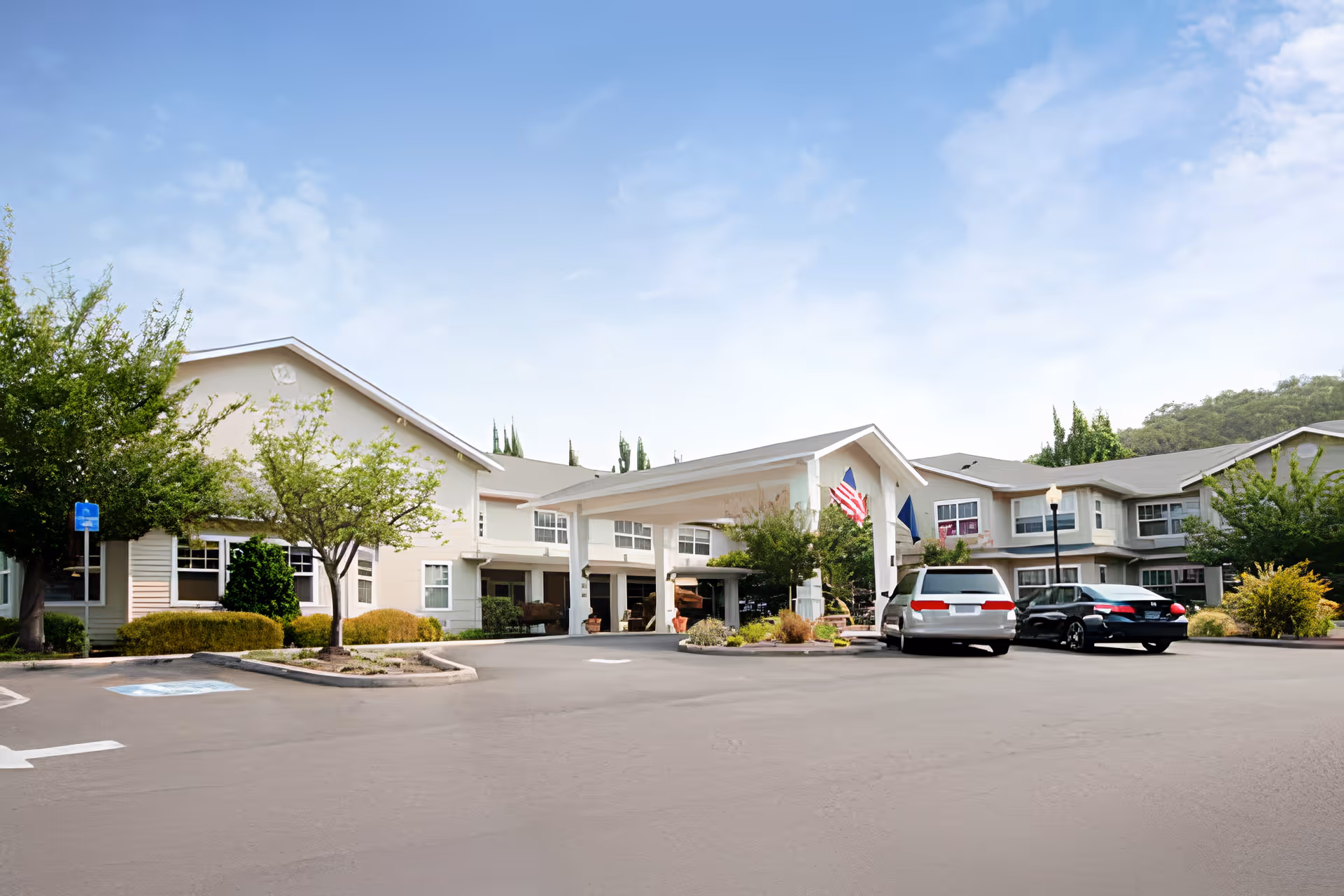 Exterior view of Oak Park Senior Living by Cogir showing a two-story building with a covered entrance, several windows, and a parking lot with cars. Trees and shrubs surround the building under a partly cloudy sky.