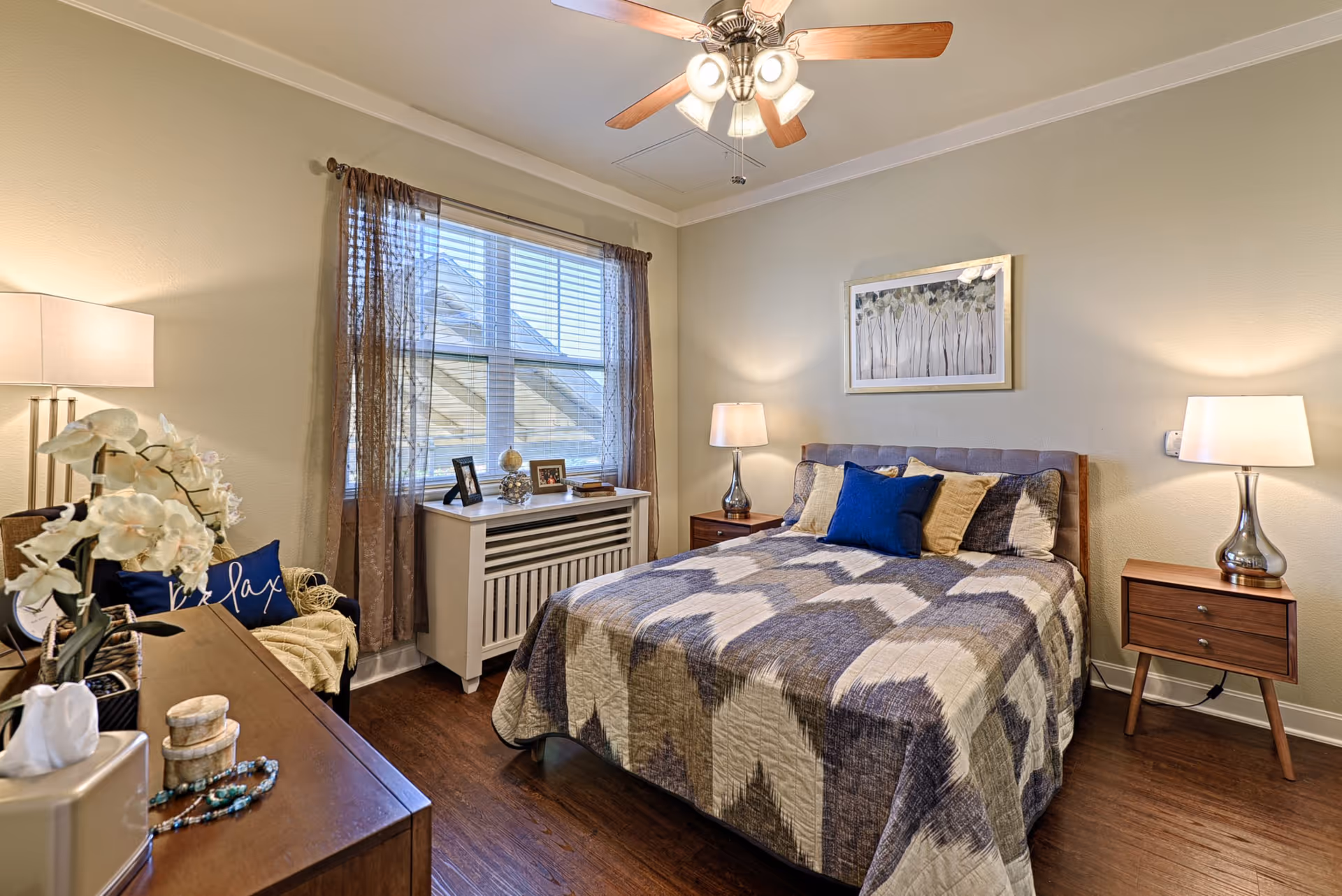 Neatly decorated bedroom with a bed, two bedside tables and lamps, a dresser, and a window with curtains.