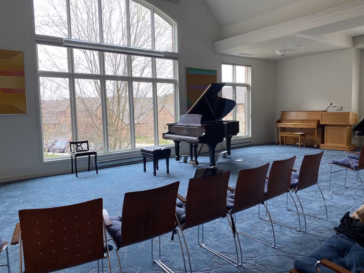 Community room with a grand piano in front of large arched windows and rows of chairs.