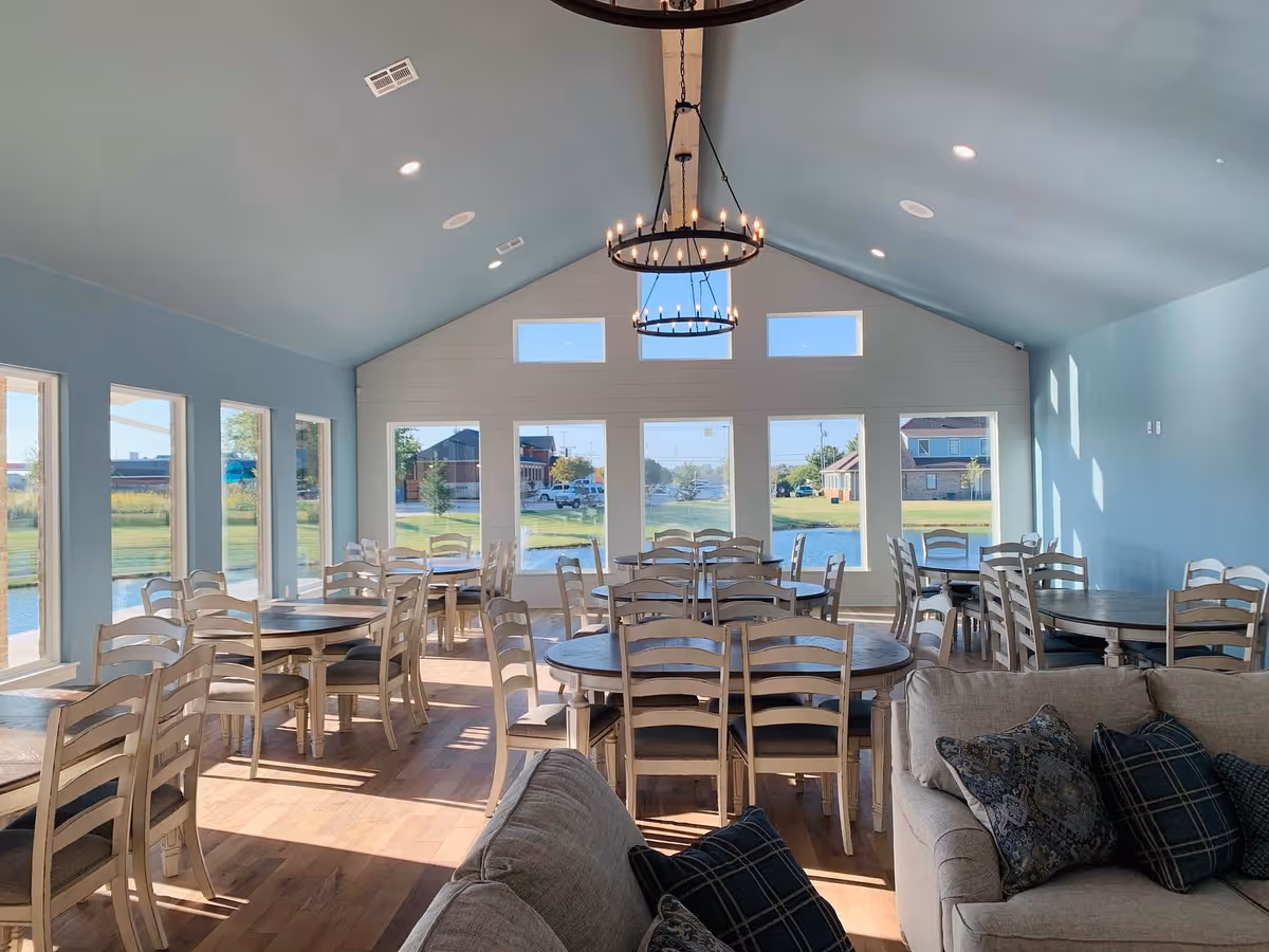 Bright communal dining room with multiple round tables and chairs, large windows overlooking a lawn and pond, and a vaulted ceiling with chandeliers.