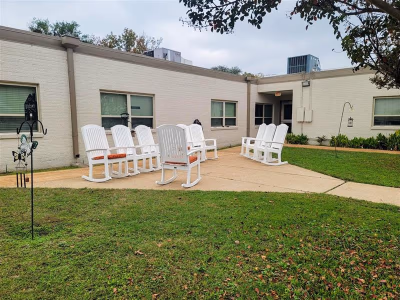 Outdoor courtyard area at Gilmer Nursing & Rehabilitation featuring a concrete patio with white rocking chairs arranged in two groups, surrounded by green grass and a light-colored brick building with windows in the background.