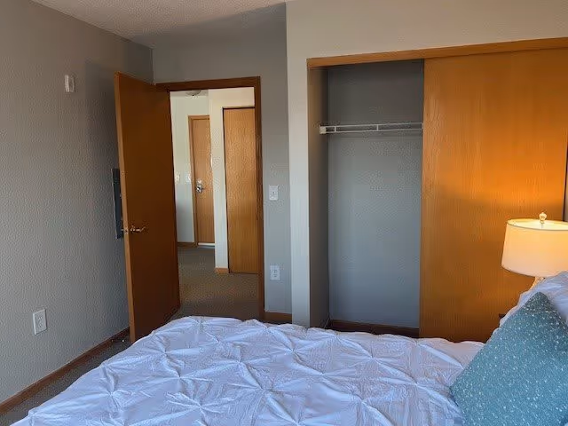 A bedroom in a senior living facility featuring a bed with a white comforter and a blue pillow, a wooden closet with sliding doors, a lamp on a nightstand, and an open door leading to a hallway with additional doors.
