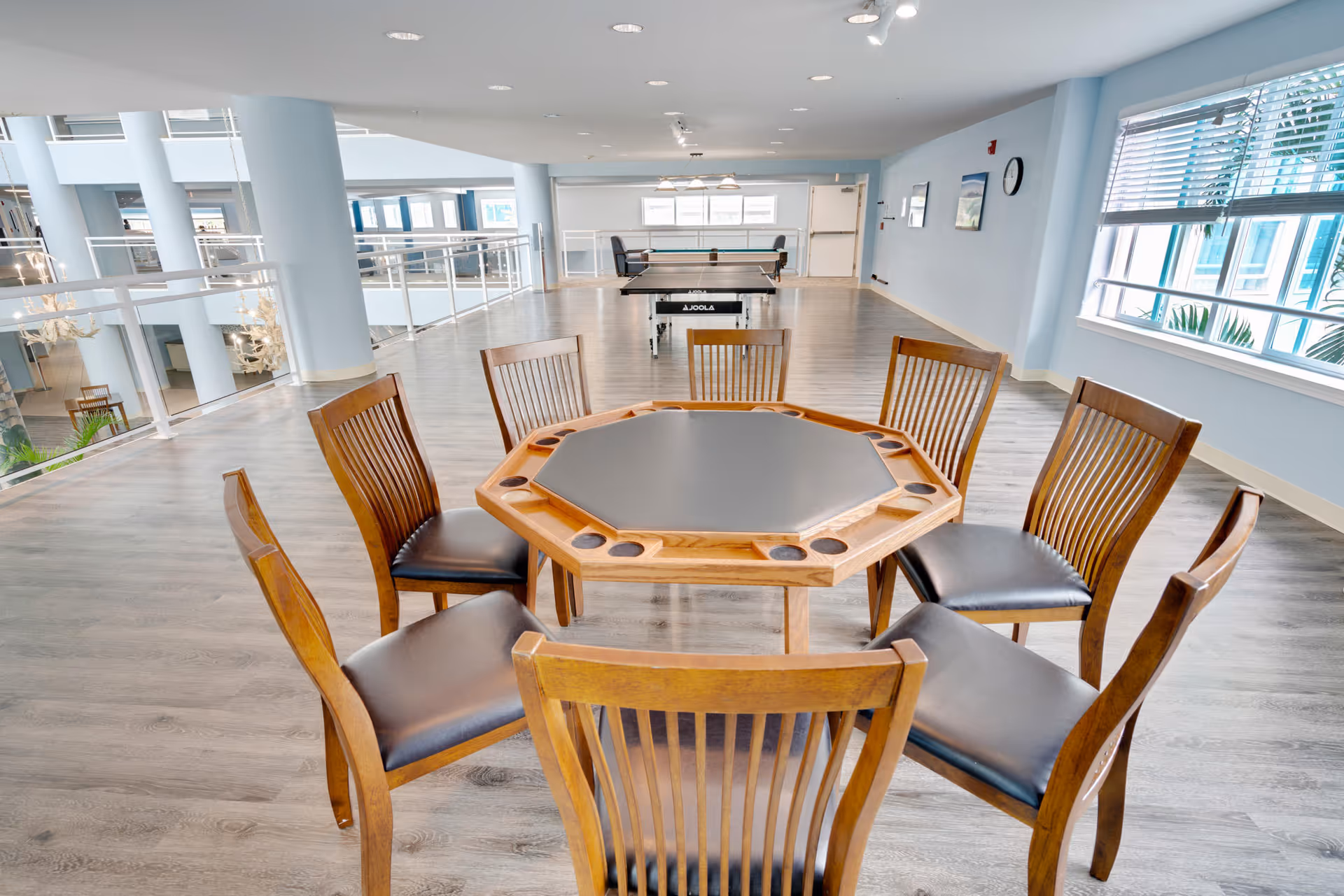 A bright, spacious common area with a wooden octagonal table surrounded by eight wooden chairs with black cushions. In the background, there is a ping pong table and large windows letting in natural light. The room has light blue walls and wood flooring.