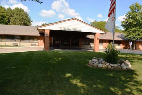 Front exterior view of East Lake Nursing and Rehabilitation Center showing a single-story building with a covered entrance, a well-maintained lawn, a rock-bordered flower bed with a flagpole, and trees in the background under a partly cloudy sky.