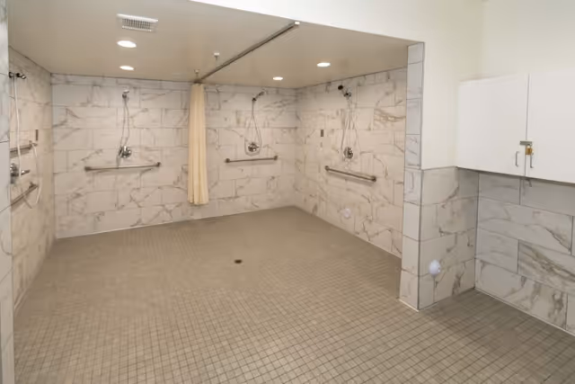 Large accessible communal shower room with multiple wall-mounted showerheads, grab bars, tiled walls and a central floor drain.