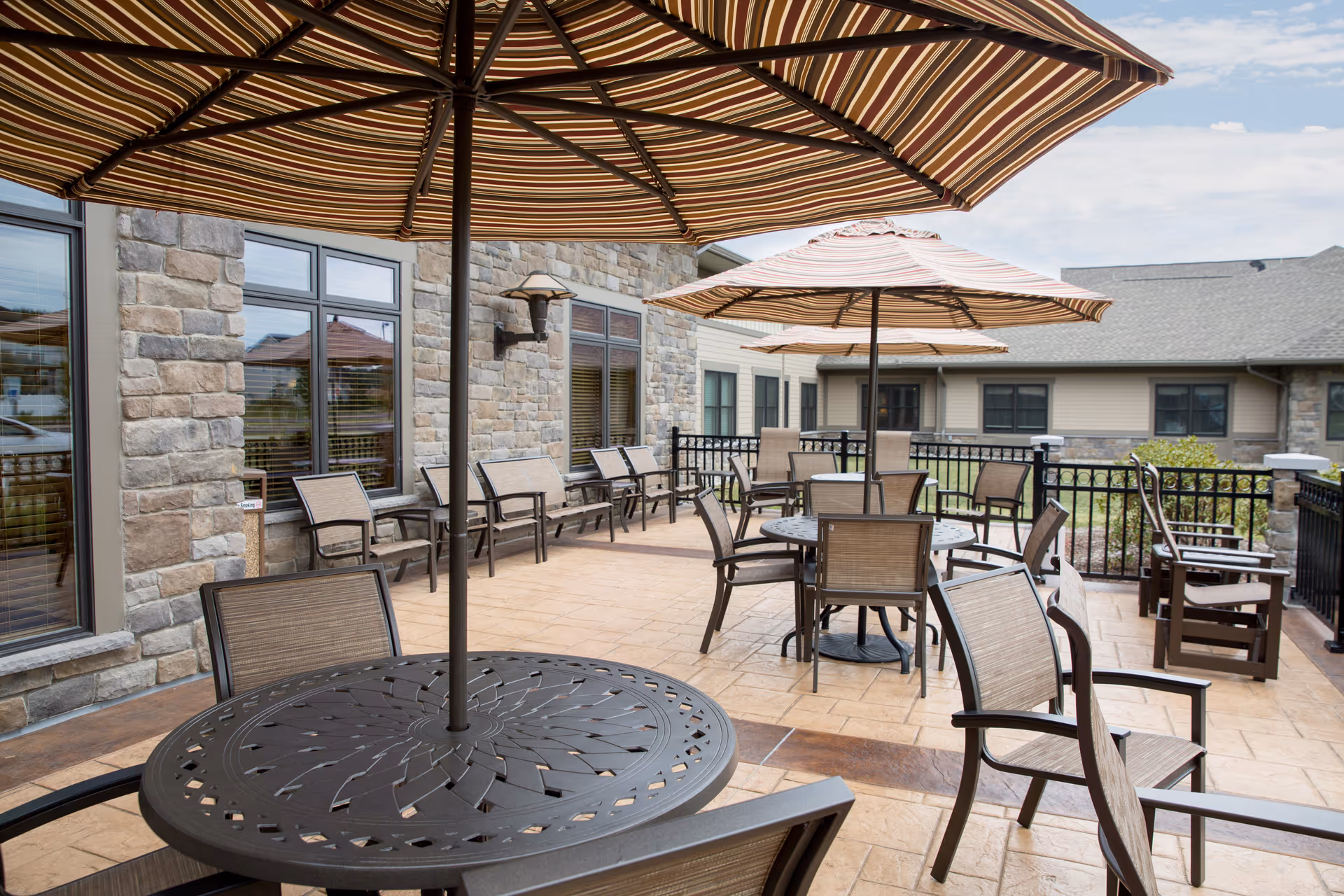 Outdoor patio area with multiple round tables and chairs, each table shaded by large striped umbrellas. The patio is paved with stone tiles and bordered by a black metal railing. The building exterior features stone walls and multiple windows.