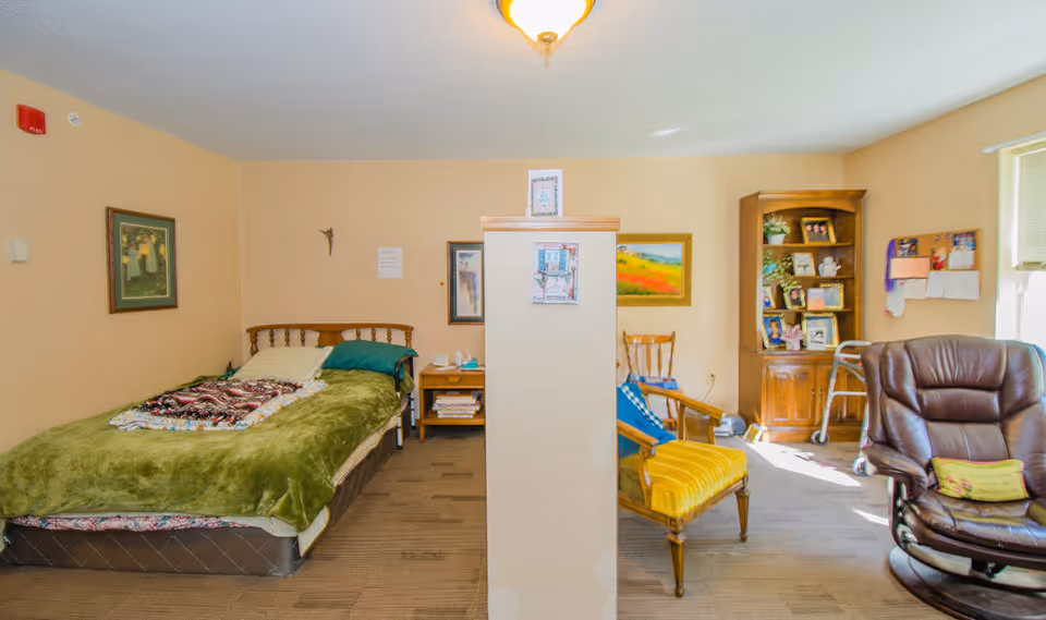 A cozy room divided by a partial wall, featuring a bed with green bedding and a blanket on the left side, and a sitting area with a yellow cushioned chair, a brown leather recliner, a wooden bookshelf with framed photos and decorations, and a walker near the window on the right side. The walls are light beige with framed artwork and a bulletin board.
