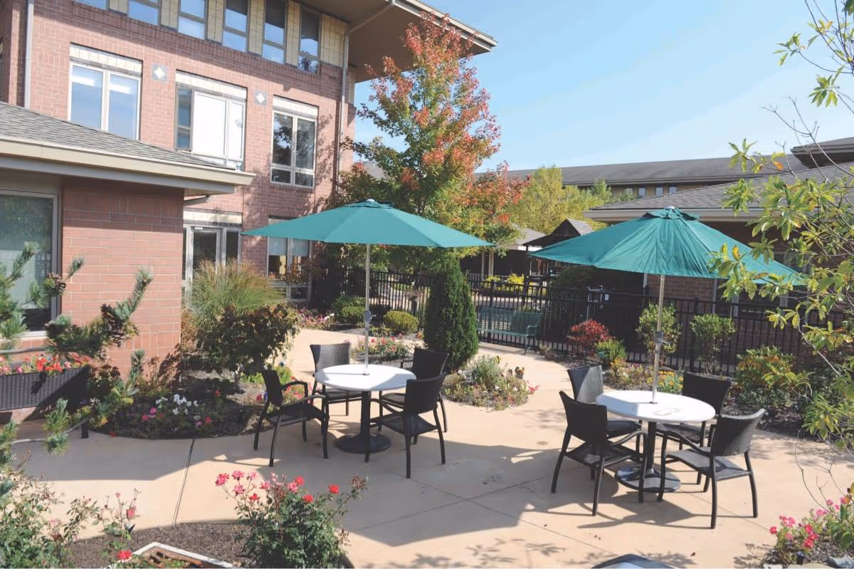 Outdoor patio area at a nursing and rehabilitation center with round tables, black chairs, and green umbrellas. The patio is surrounded by landscaped flower beds and trees with a multi-story brick building in the background.