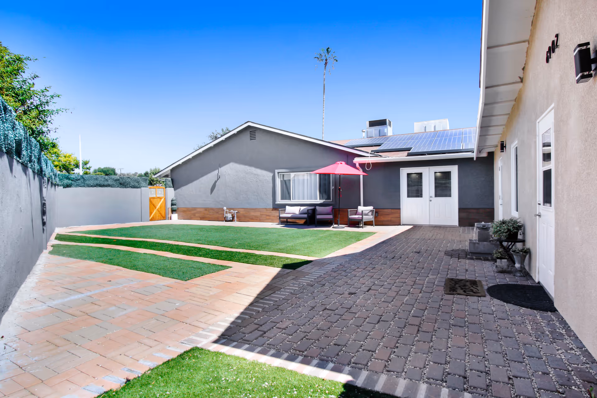 Outdoor courtyard area of a retirement residence with a paved walkway, artificial grass patches, a gray building with solar panels on the roof, white double doors, and outdoor seating under a red umbrella.