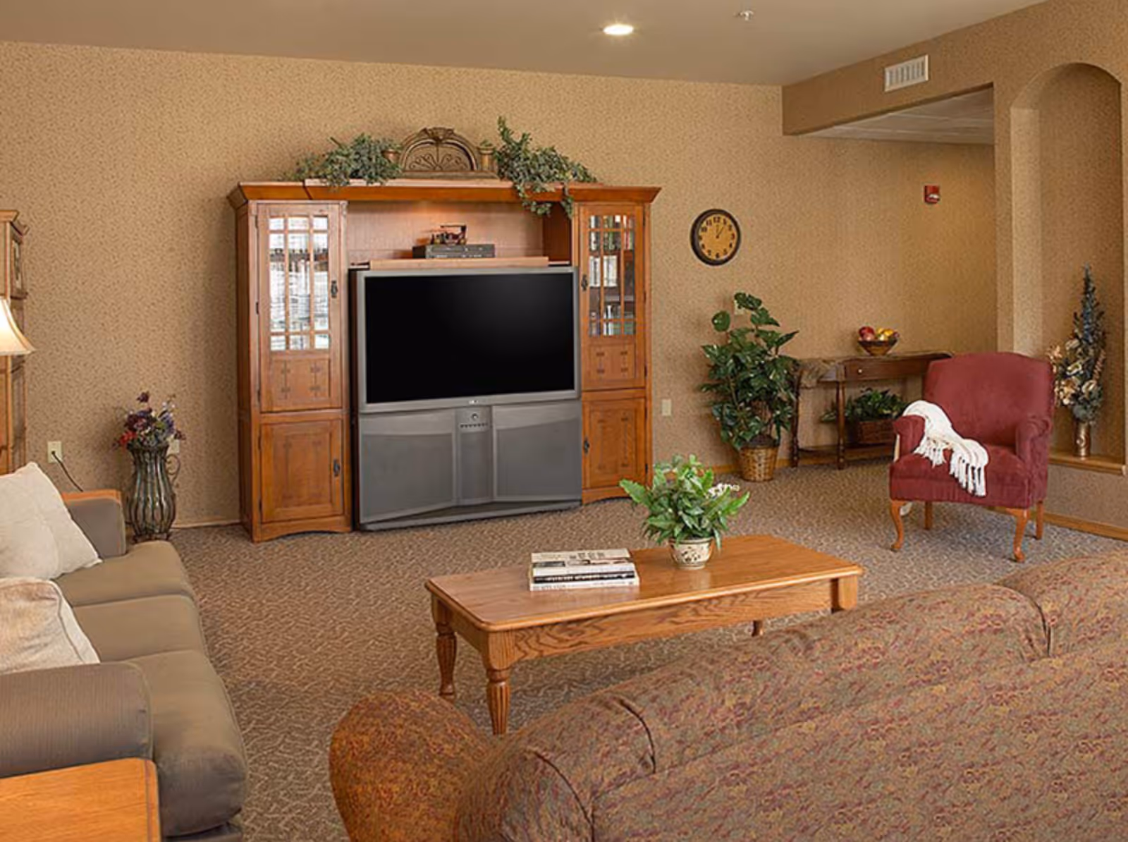 A cozy living room with a large wooden entertainment center housing a flat-screen TV. The room features a beige sofa, a patterned couch, a wooden coffee table with a plant and books, a red armchair with a white throw, and various plants and decorative items. The walls are beige with a subtle pattern, and there is a round clock on the wall.
