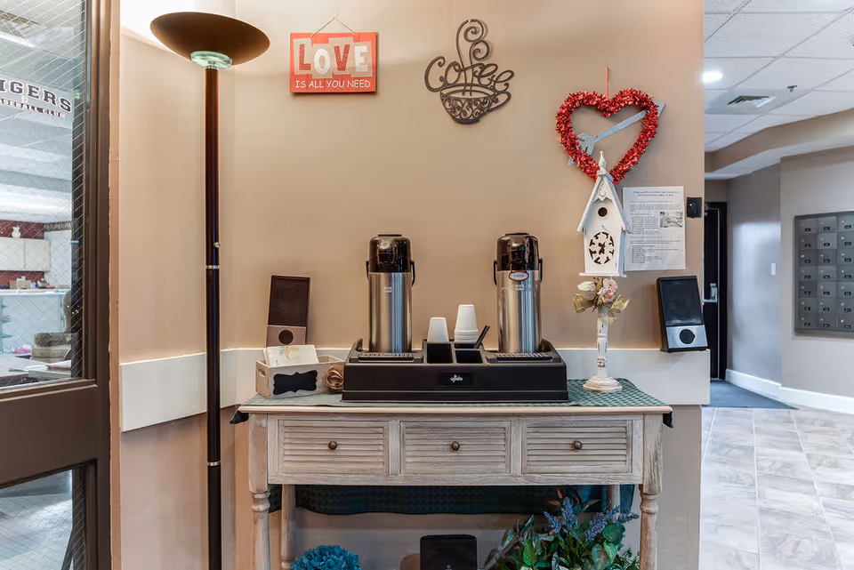 A coffee station setup in a hallway corner with two large coffee dispensers, disposable cups, and a decorative birdhouse on a wooden table. The wall behind has a red sign that says 'LOVE IS ALL YOU NEED', a metal coffee cup decoration, and a heart-shaped wreath. A tall floor lamp stands to the left, and a door with a window is partially visible on the left side.
