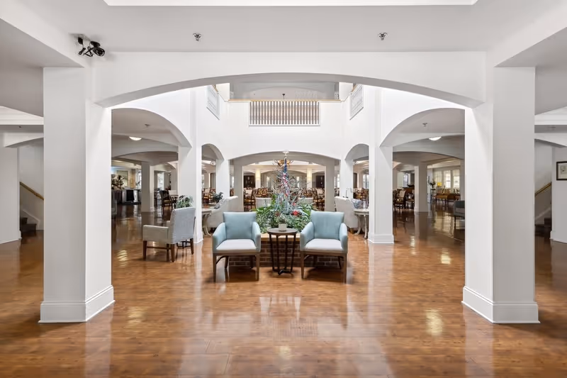 Spacious and bright common area in a senior living facility with wooden floors, white walls, and large archways. The room features comfortable seating including light blue armchairs and a small table with plants in the center. In the background, there are multiple dining tables and chairs arranged neatly.