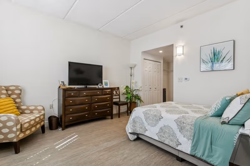 A bedroom with a bed covered in a patterned white and gray bedspread with teal and white pillows. To the left is a wooden dresser with a flat-screen TV on top, a chair, a floor lamp, and a patterned armchair with a yellow pillow. There is a framed plant artwork on the wall and a closet with double doors in the background.