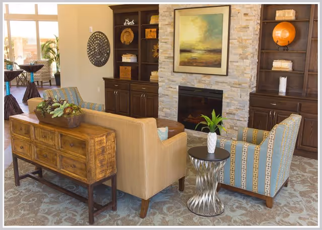 Bright furnished common living area with sofas and armchairs arranged around a stone fireplace flanked by built-in shelving and decorative accents.