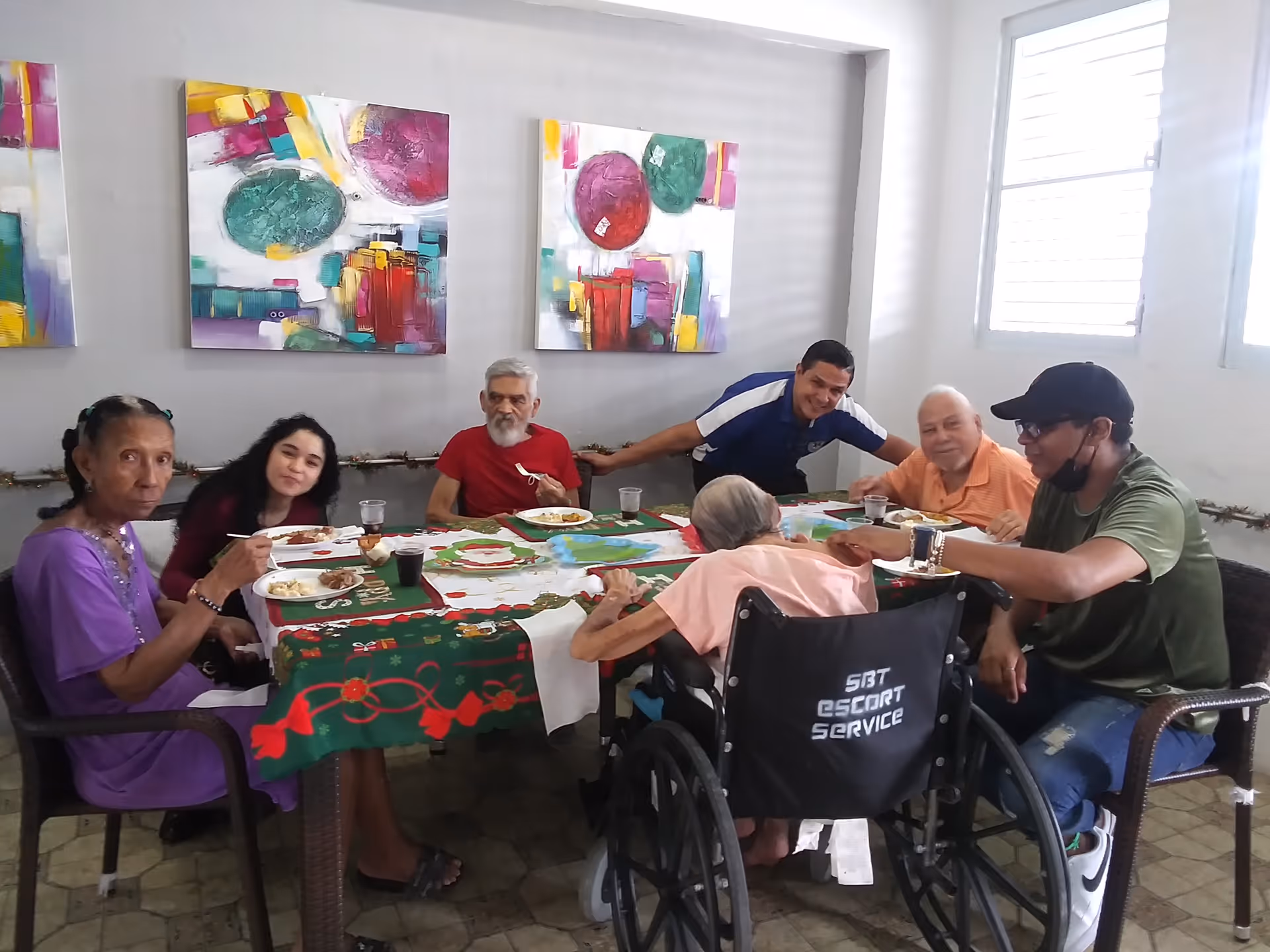 Elderly residents and caregivers sit around a decorated dining table sharing a meal in a bright common room.