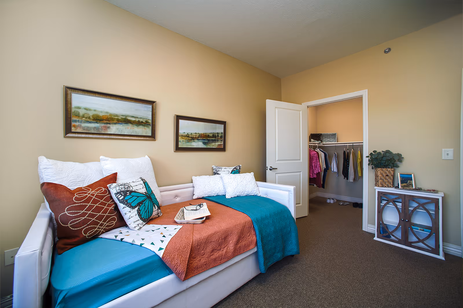 Bright bedroom with a daybed dressed in colorful pillows and throws, an open closet, and a small cabinet.