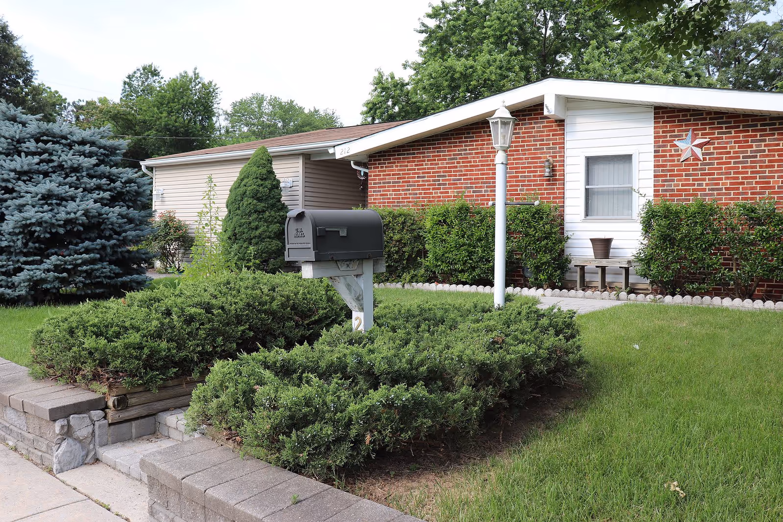 Front yard of a single-story brick and siding house with a black mailbox on a white post surrounded by green bushes, a white lamp post, a small bench with a flower pot, and a decorative star on the house wall. The yard is well-maintained with green grass and various shrubs and trees.