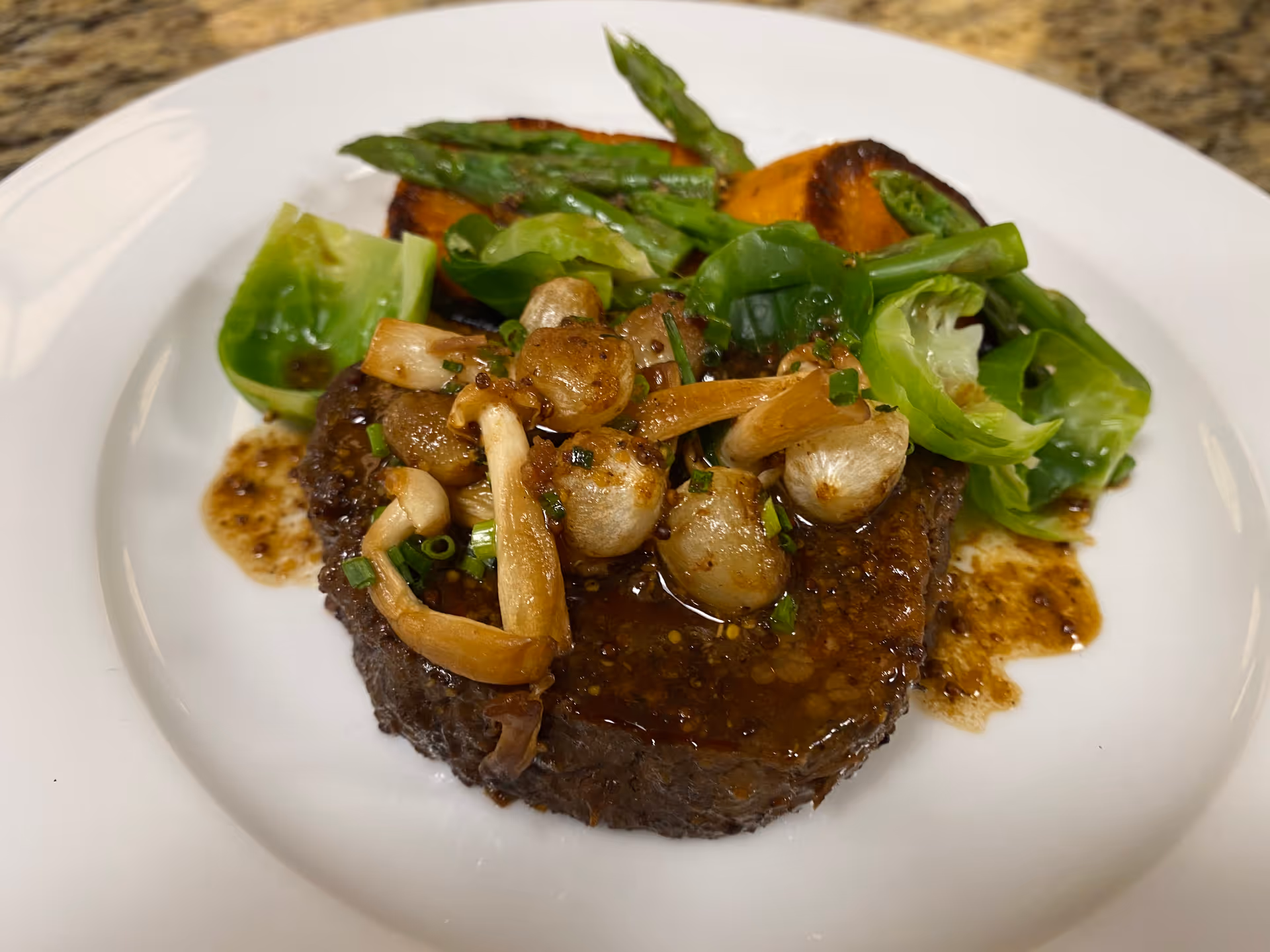 A plated meal featuring a cooked piece of meat topped with sautéed mushrooms and herbs, accompanied by roasted sweet potatoes, asparagus, and Brussels sprouts on a white plate.