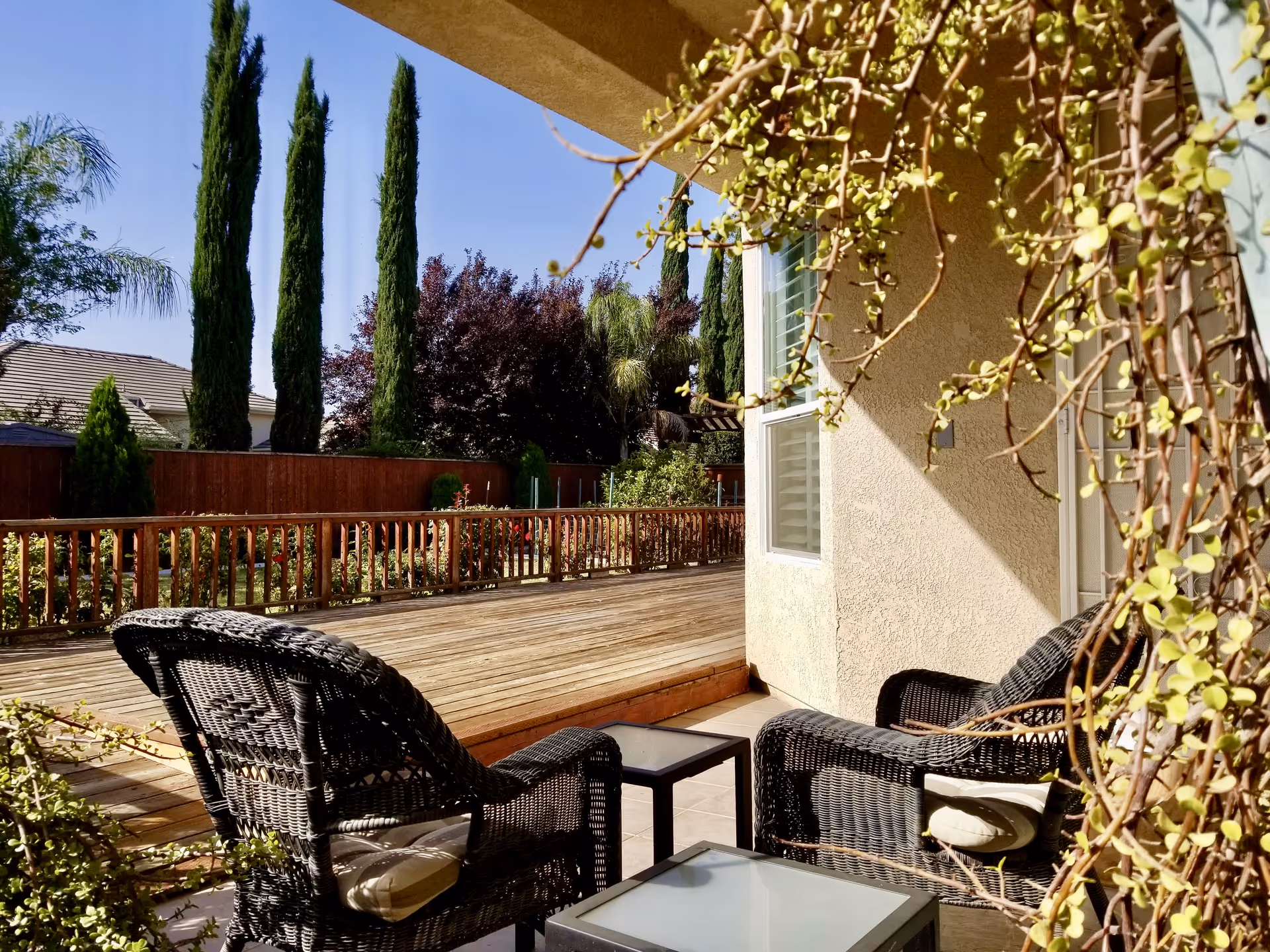 Outdoor patio area with two black wicker chairs and two small glass-top tables. The patio overlooks a wooden deck and a garden with tall trees and various plants under a clear blue sky.