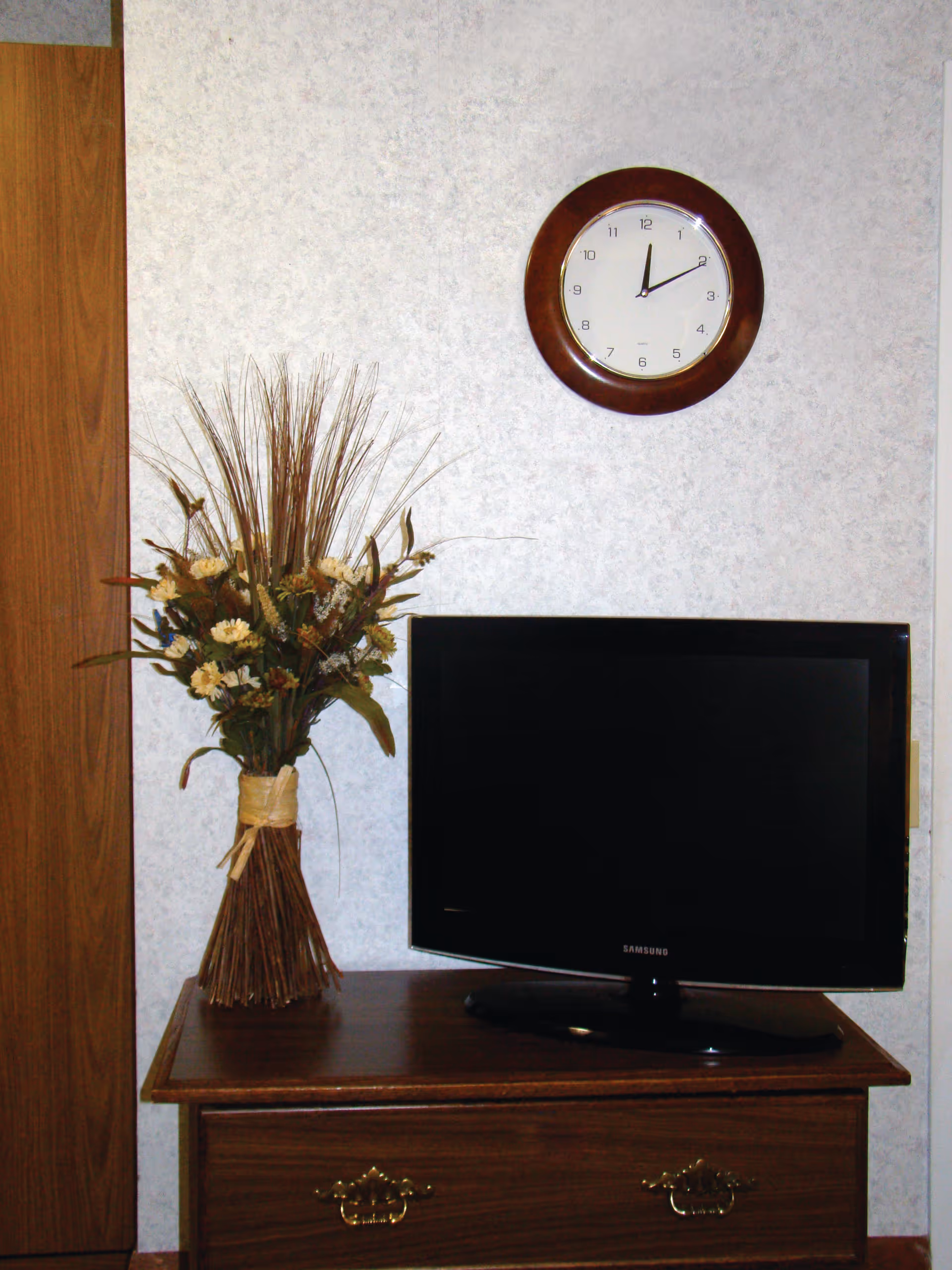 A wall clock hangs above a wooden dresser holding a flat-screen TV and a vase of dried flowers.