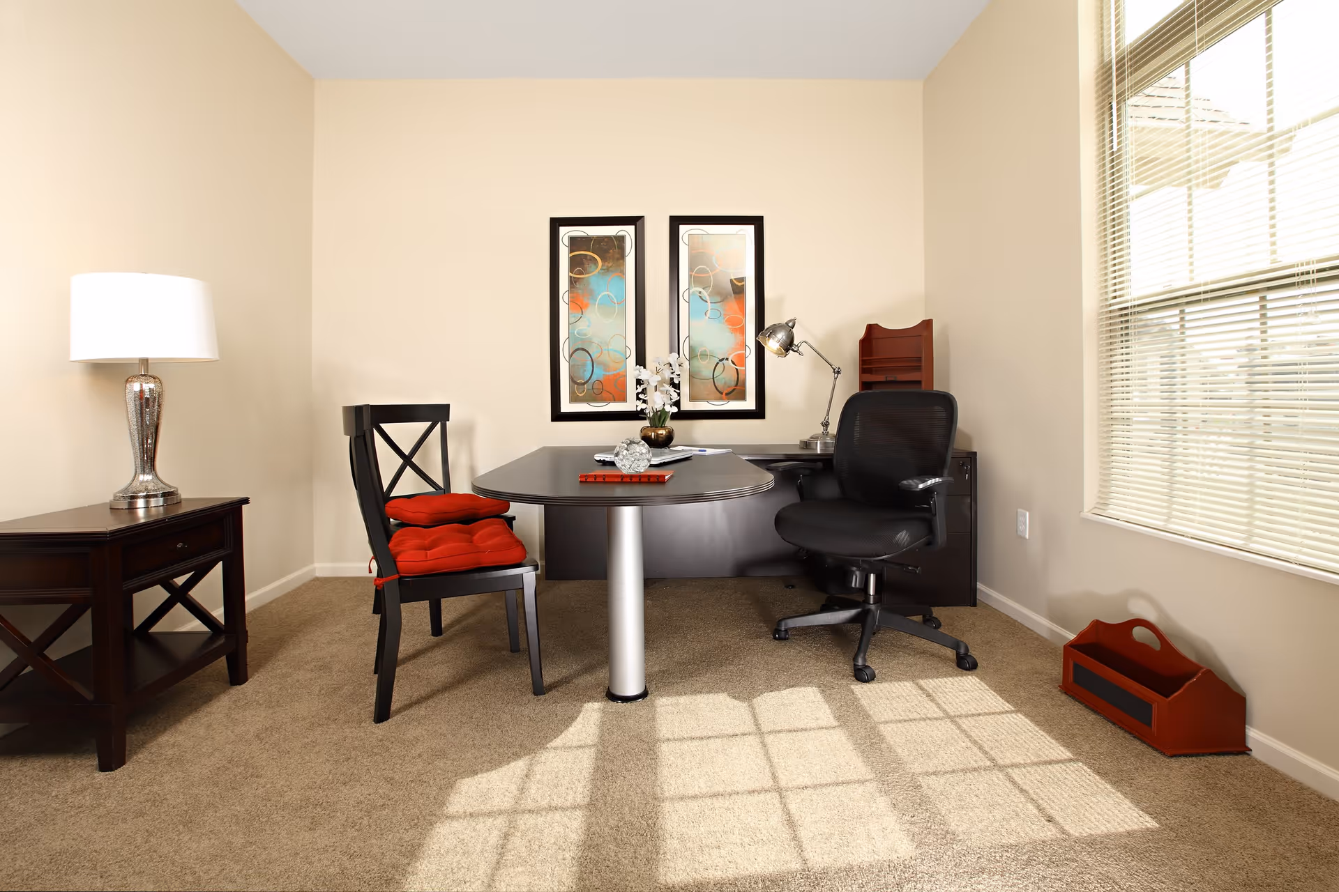 A small office space with a dark wooden desk and two chairs, one with red cushions and the other a black swivel chair. There is a table lamp on a side table to the left, two abstract framed artworks on the wall, and a window with blinds on the right letting in sunlight that casts shadows on the carpeted floor.