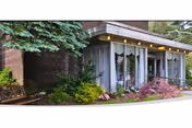 Low brick building entrance with large glass windows, an overhang with lights, and landscaped shrubs and a bench.