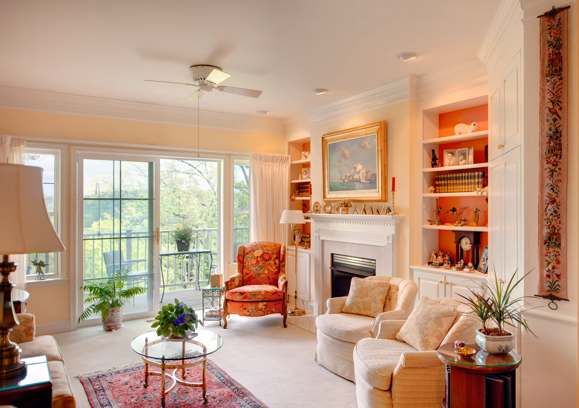 A cozy living room with a large sliding glass door leading to a balcony with outdoor chairs and a table. Inside, there is a floral armchair, two cushioned chairs with pillows, a glass coffee table with a plant, a fireplace with a painting above it, built-in shelves with books and decorative items, and a ceiling fan. The room is warmly lit and decorated with plants and a patterned rug.