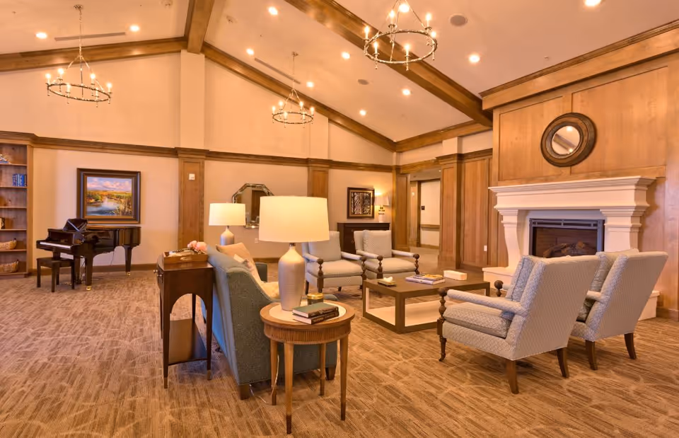 Warm, spacious communal living room with upholstered seating, a fireplace, piano, and vaulted wood-beamed ceiling.