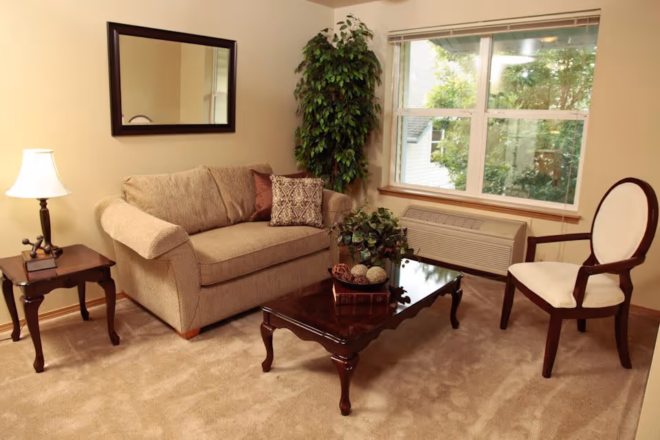 A cozy living room with a beige loveseat adorned with two decorative pillows, a wooden side table with a lamp and a small sculpture, a wooden coffee table with decorative items and a book, a white cushioned wooden chair, a large window with a view of greenery outside, a wall-mounted mirror, and a tall potted plant in the corner.