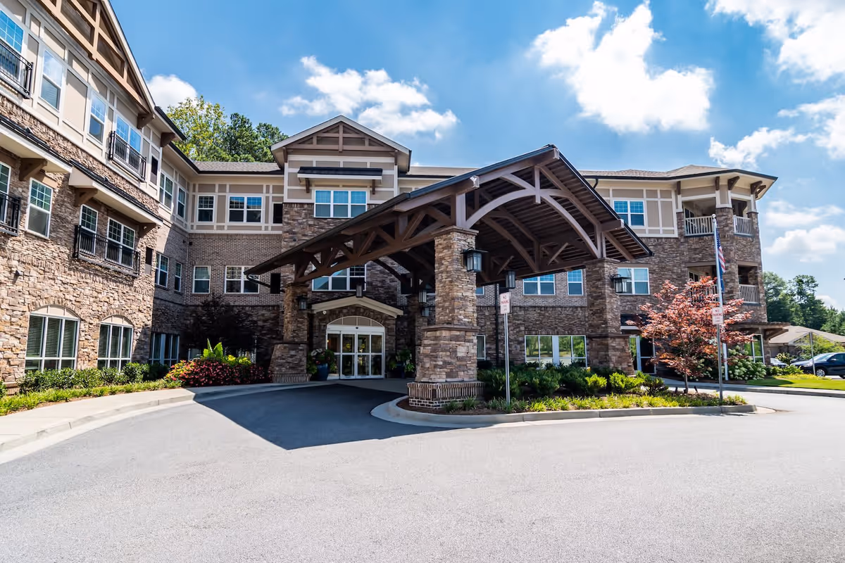 Front exterior view of Arbor Terrace Burnt Hickory, a multi-story senior living facility with stone and brick facade, large covered entrance with wooden beams, surrounded by landscaped greenery and a clear blue sky with some clouds.