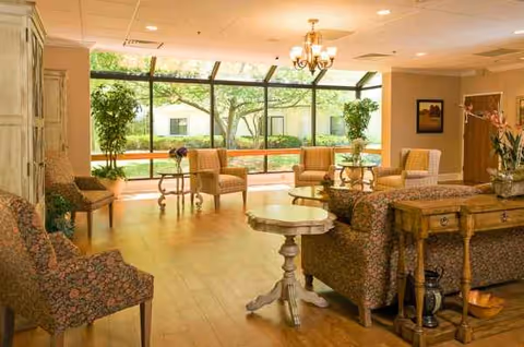 Bright senior living lounge with chairs, tables, plants, a chandelier, and large sunny windows.
