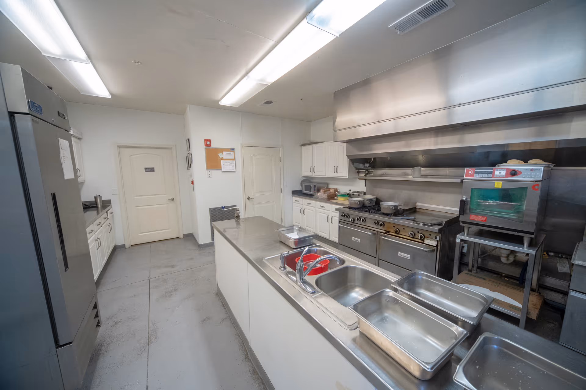A commercial kitchen with stainless steel appliances including a large refrigerator, stove, oven, and multiple sinks. White cabinets line the walls and there are fluorescent lights on the ceiling. Various kitchen utensils and containers are placed on the countertops.