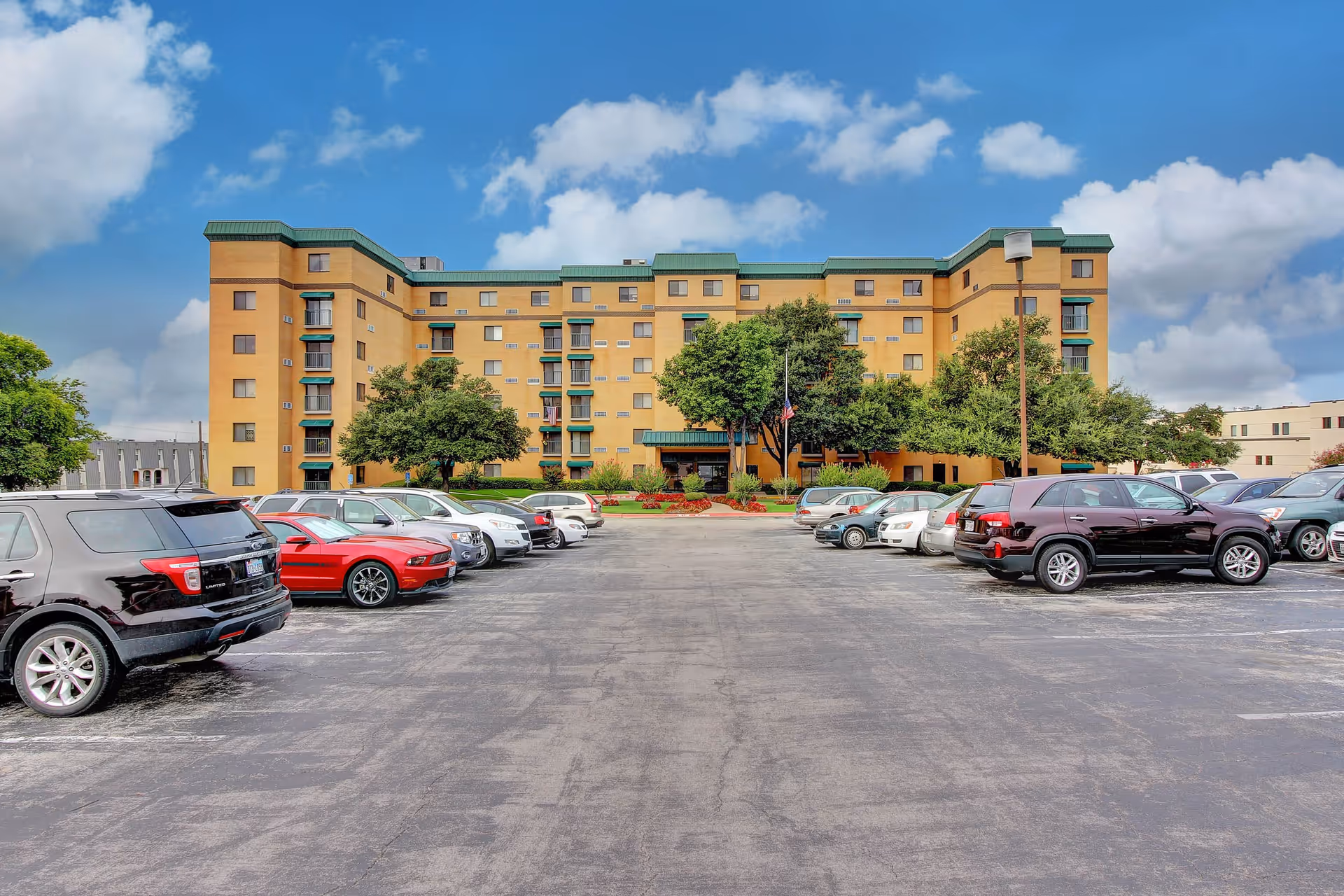 Front exterior view of a multi-story yellow building with green roof accents, surrounded by trees and a parking lot filled with cars under a partly cloudy blue sky.