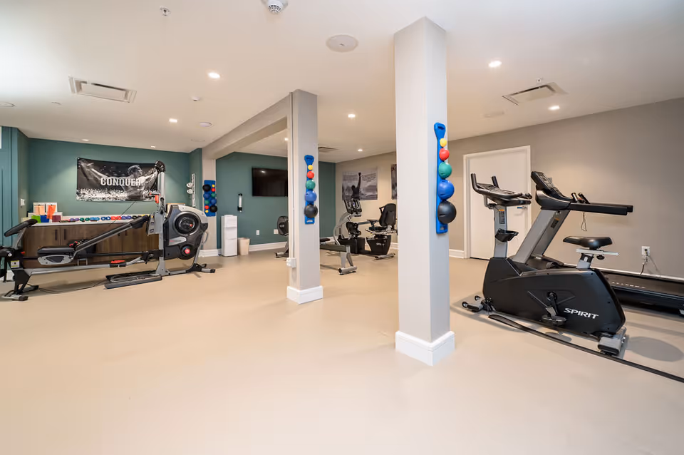 Bright fitness room with exercise equipment including treadmills, stationary bikes, and a rowing machine arranged around support pillars.