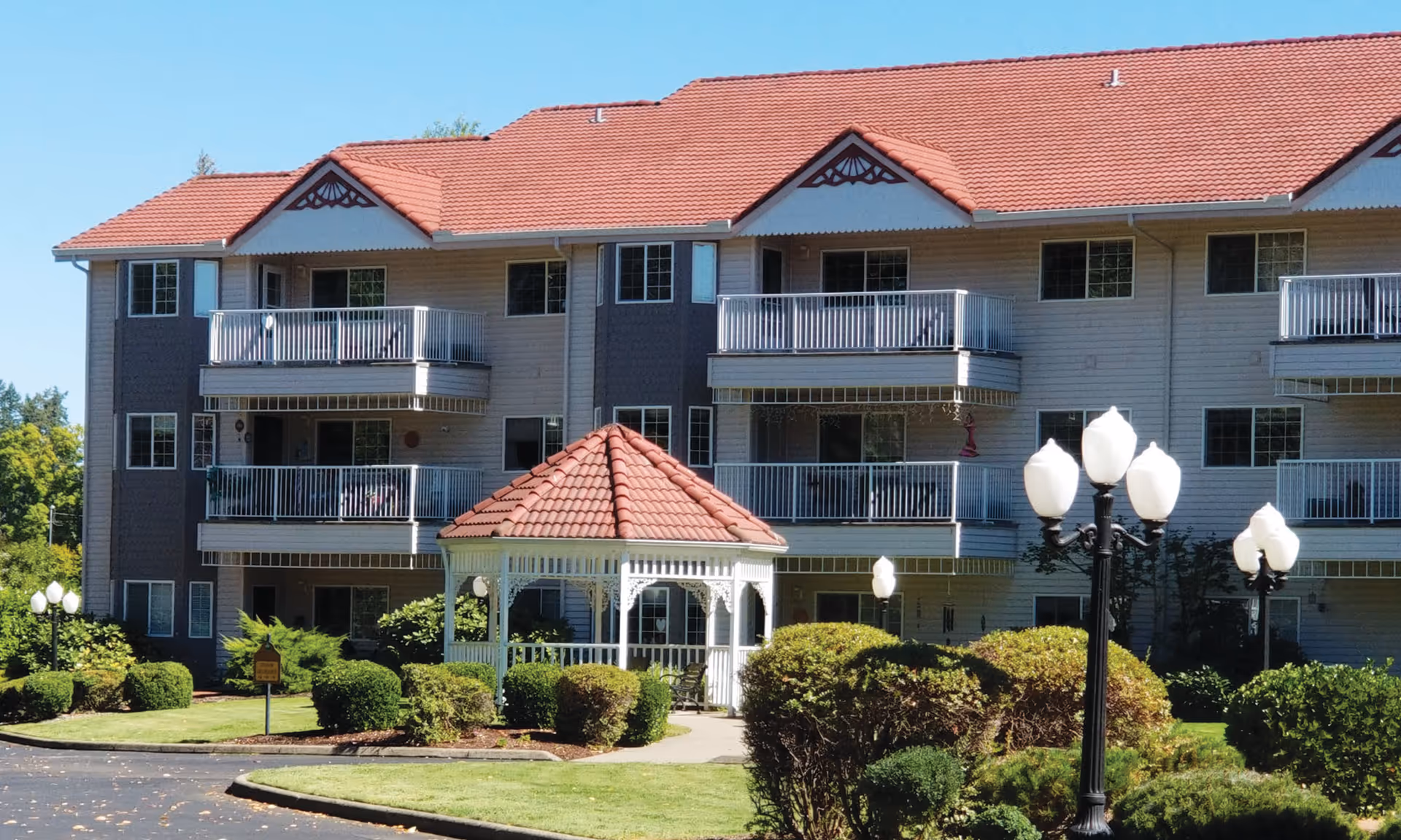 Exterior view of a multi-story residential building with beige siding and red tiled roofs. The building features multiple balconies with white railings. In front of the building is a well-maintained garden with trimmed bushes, a white gazebo with a red tiled roof, and several vintage-style street lamps with white globes. The sky is clear and blue.