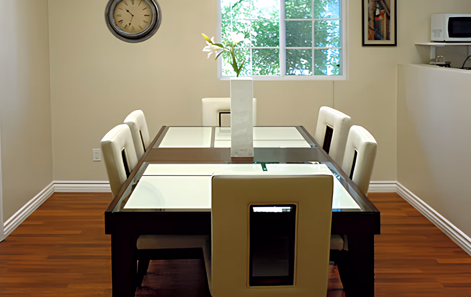 A dining room with a rectangular glass-top table surrounded by six white cushioned chairs. A tall white vase with flowers is placed in the center of the table. There is a window with greenery visible outside, a wall clock, and a microwave on a counter in the background.