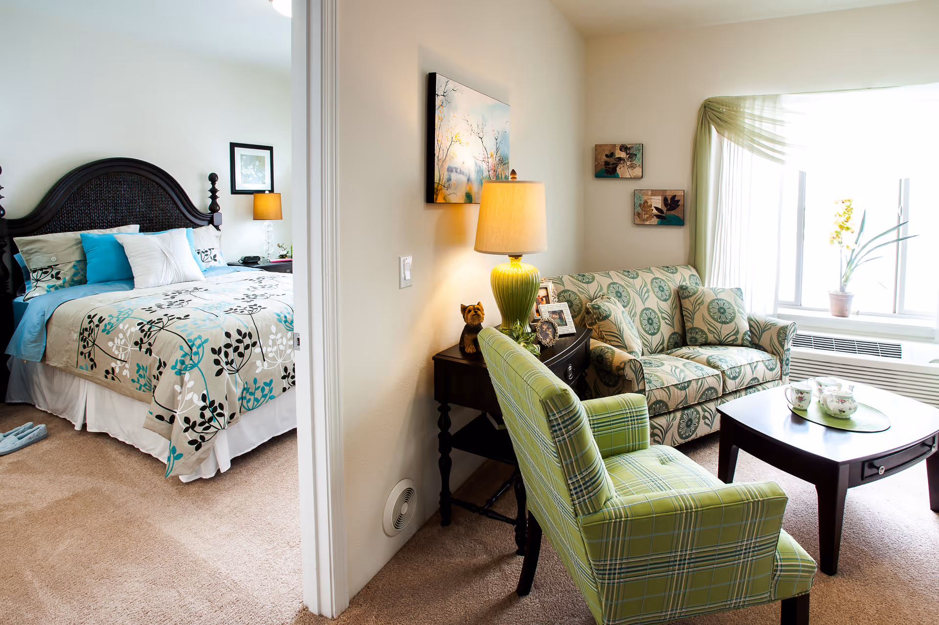 A cozy living area with a green patterned armchair, a floral patterned loveseat, a dark wooden coffee table with a tea set, and a side table with a green lamp and decorative items. Adjacent to the living area is a bedroom with a bed featuring a floral bedspread in shades of beige, turquoise, and black, with multiple pillows and a bedside table with a lamp.