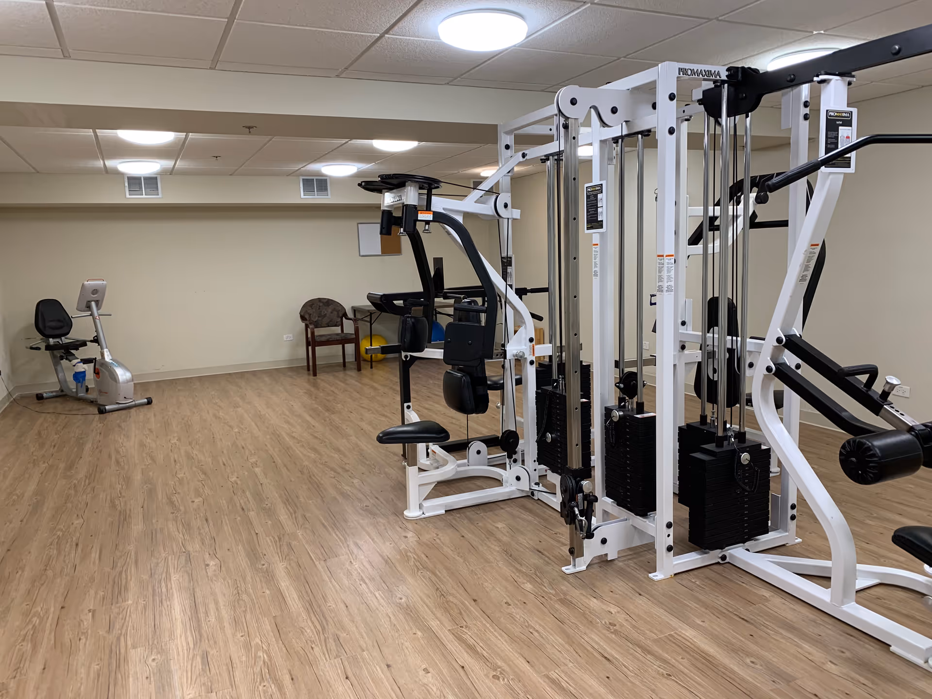 Indoor fitness room with exercise equipment including a recumbent bike, a multi-station weight machine, and a chair against a beige wall. The room has wood-patterned flooring and ceiling lights.