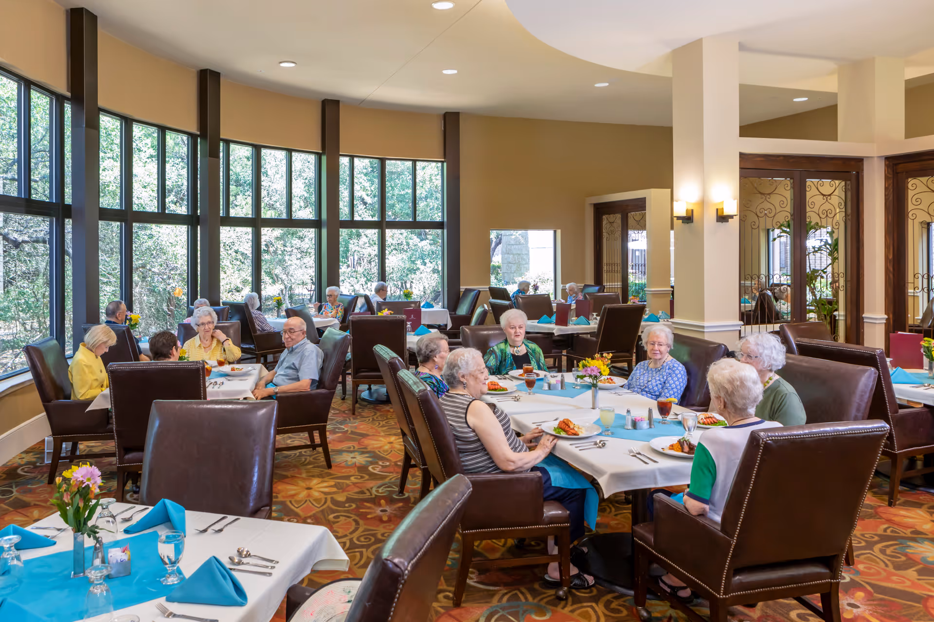 Several elderly residents seated at tables in a bright, spacious dining room with large windows.