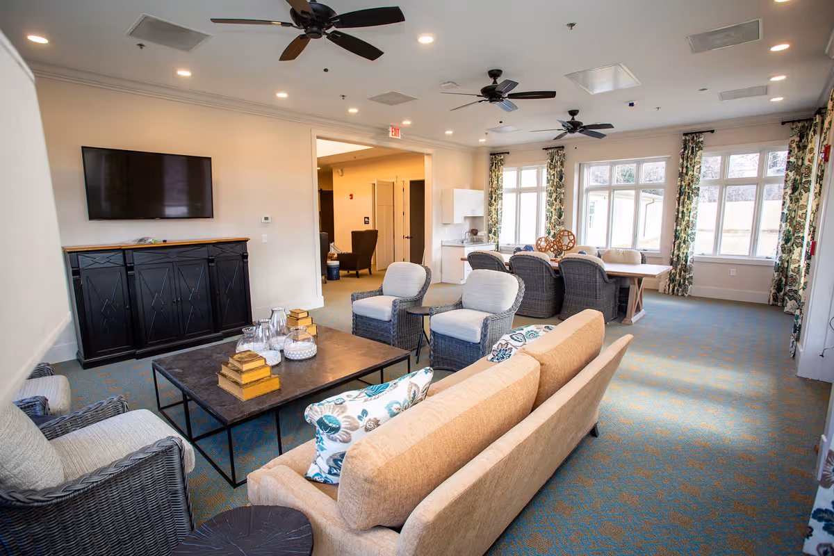 A bright and spacious senior living common area with beige and gray cushioned chairs, a beige sofa with floral pillows, a large coffee table with decorative items, a flat-screen TV mounted on the wall, ceiling fans, and large windows with floral curtains allowing natural light to fill the room.