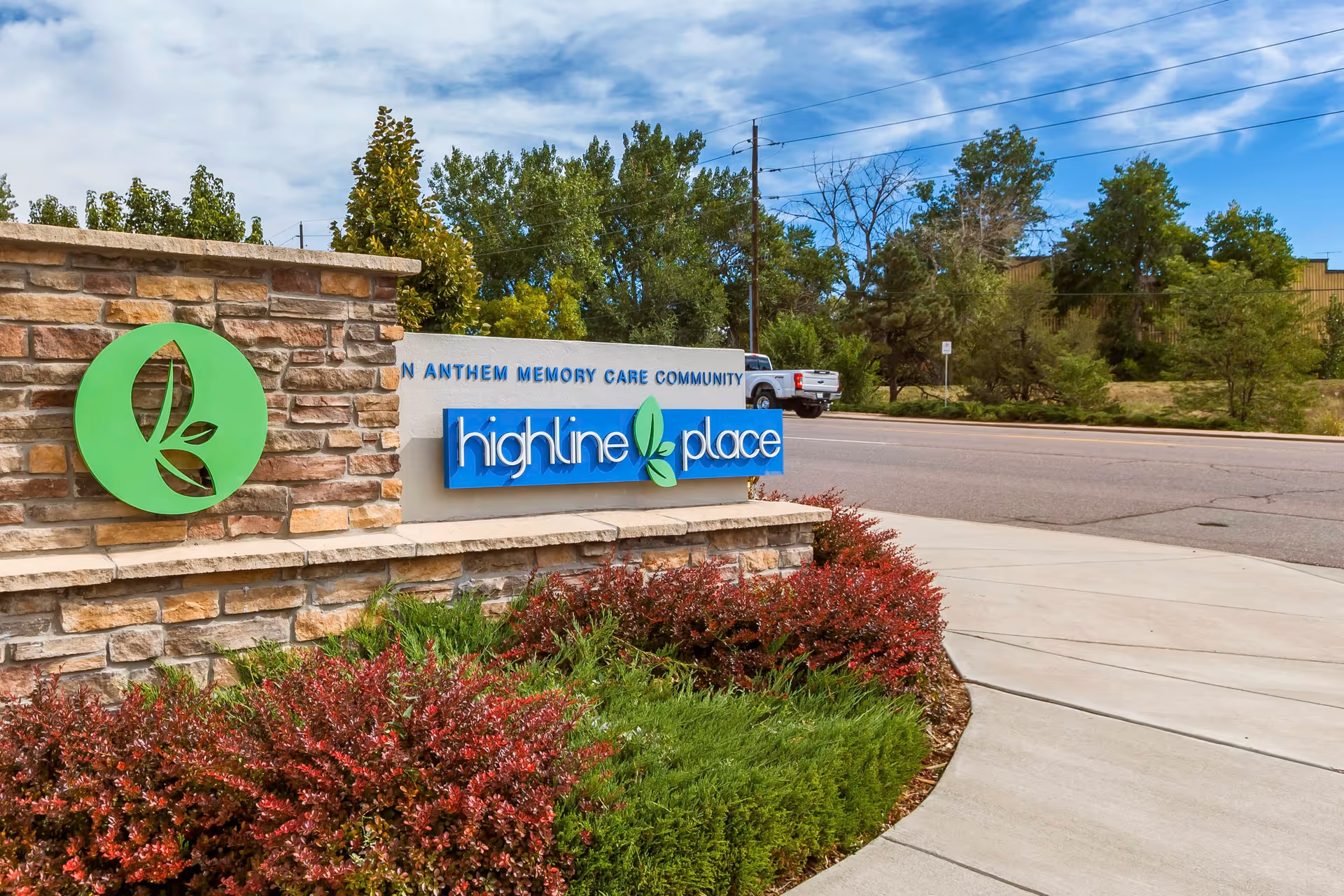 Outdoor view of a stone and concrete sign for Highline Place Memory Care Community, surrounded by green and red shrubs, with a street and trees in the background under a partly cloudy sky.