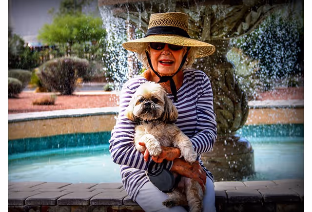 An elderly woman wearing a wide-brimmed straw hat and sunglasses sits on the edge of a stone fountain holding a small fluffy dog. The fountain is active with water cascading down, and there are bushes and landscaping in the background.