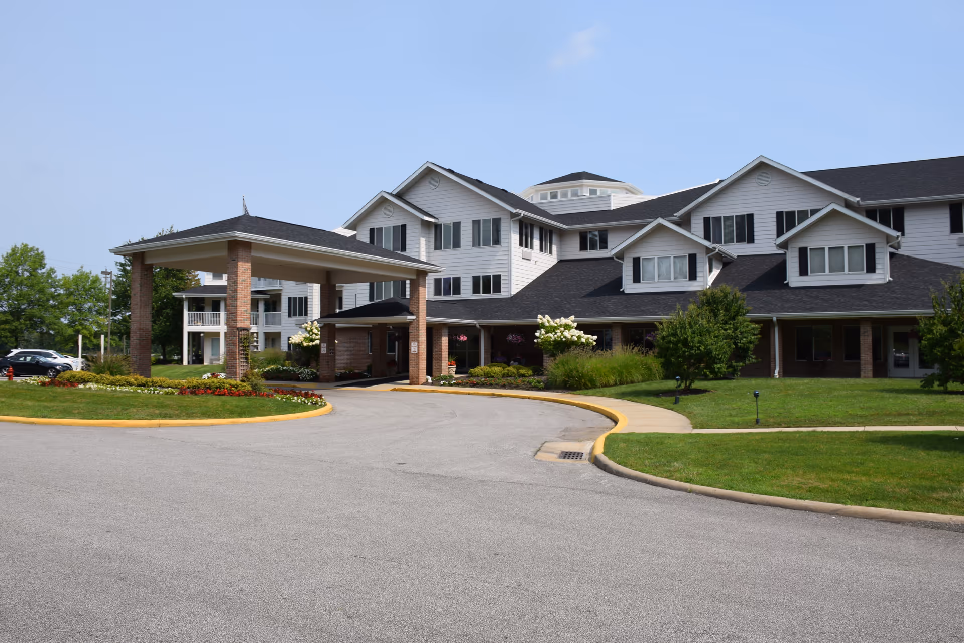 Exterior view of a large, multi-story senior living facility with white siding and black shutters. The building has a covered entrance supported by brick columns, surrounded by well-maintained landscaping with green grass, bushes, and flowers. The sky is clear and blue.