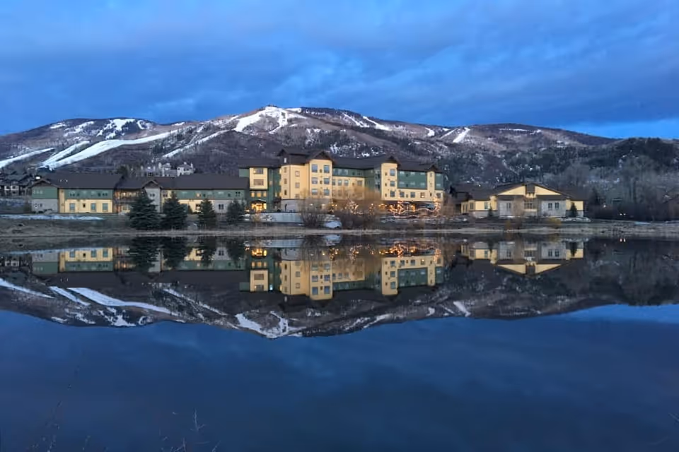 A multi-building lakeside facility reflected in a calm lake with snow-dusted mountains behind it at dusk.